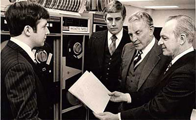 A group of men, including Ed Blount, looking over a printout from an IBM magnetic tape drive computer.